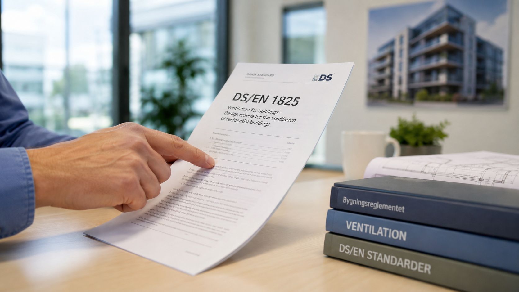 En person holder et dokument om ventilationskrav til boligbyggeri med titlen DS/EN 1825 ved et skrivebord.